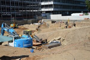 Bagger fressen sich derzeit tief in den Boden der TRON-Baugrube - im Hintergrund Archäologen, die in mehreren Teams in der Grube Funde begutachten. - Foto: gik