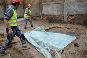 Bauarbeiter sichern römische Mauerreste in der Baugrube am Bischofsplatz mit einer Plane. - Foto: gik